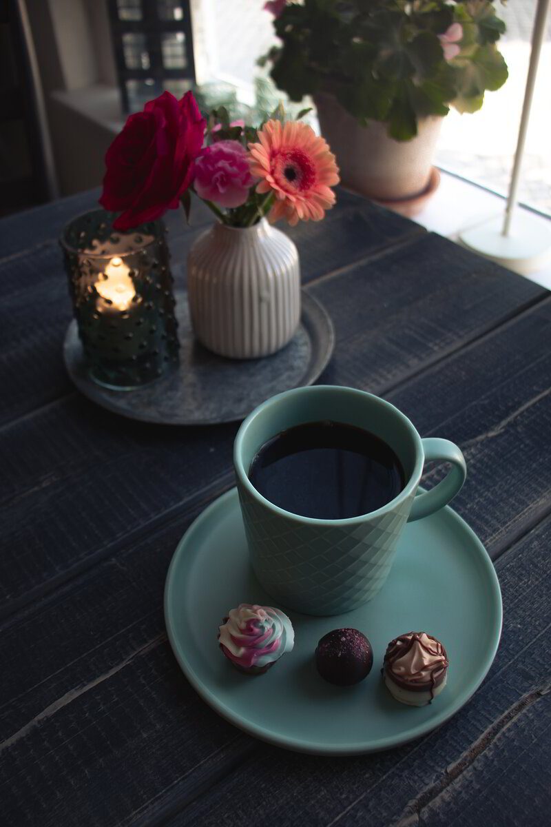 Kopp kaffe med chokladbollar på turkost fat, blommor och ljus i bakgrunden